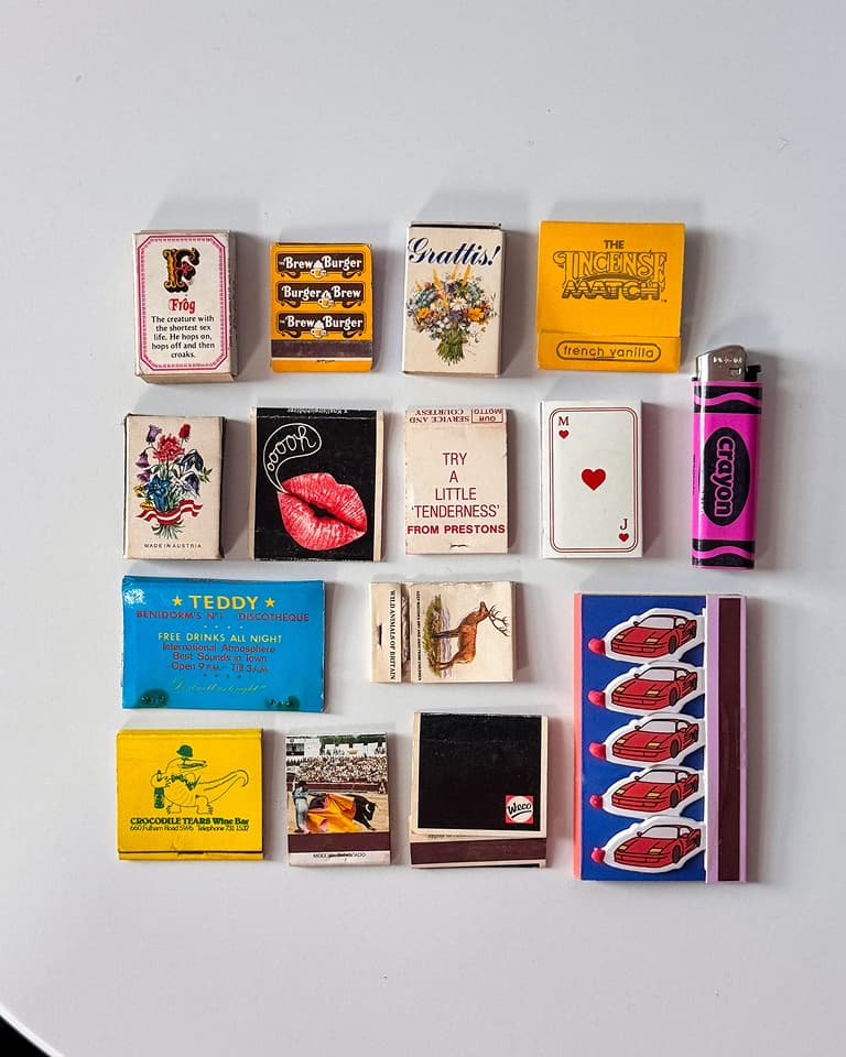 Flat lay of 16 vintage matchboxes and a pink lighter arranged in a grid on white background, featuring colorful retro logos and illustrations.