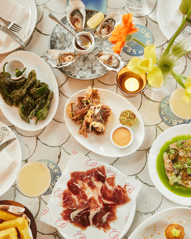 An elegant table setting with dishes including oysters on ice, roasted peppers, sliced cured meat, fried seafood, and decorative flowers.