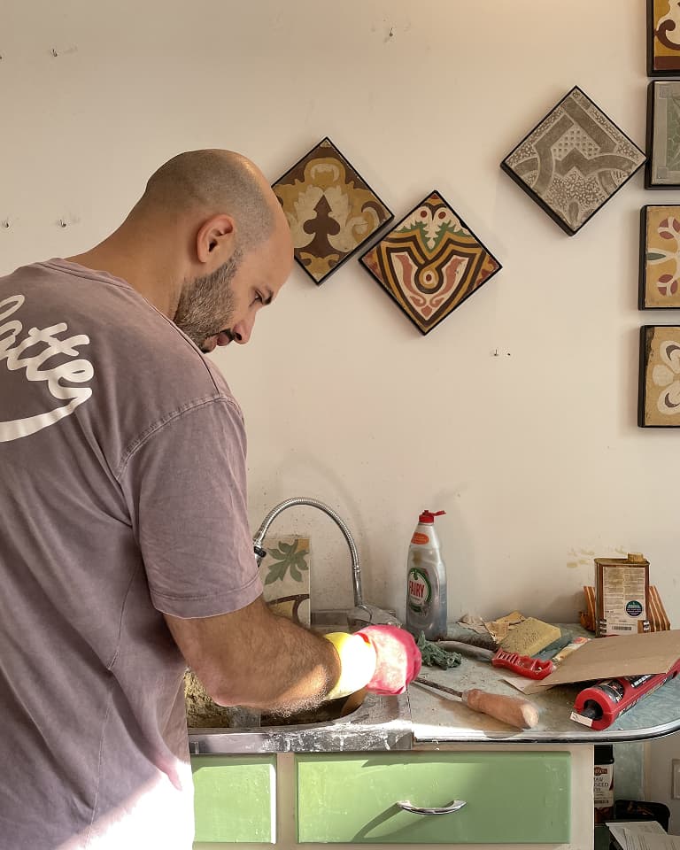 Man preparing food at a kitchen counter with tiles on the wall. Various utensils and cleaning supplies are visible on the counter.