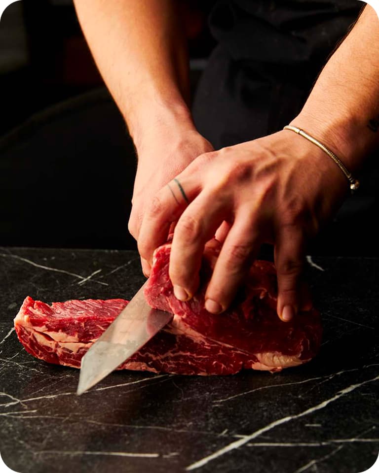 Hands slicing a raw steak with a chef's knife on a dark marble countertop.