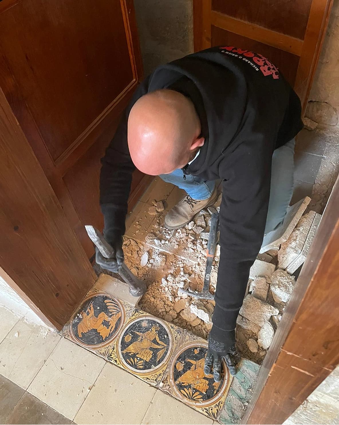 Person removing floor tiles with tools, revealing patterned tiles underneath. Wearing gloves, jeans, and a black hoodie in a small space.