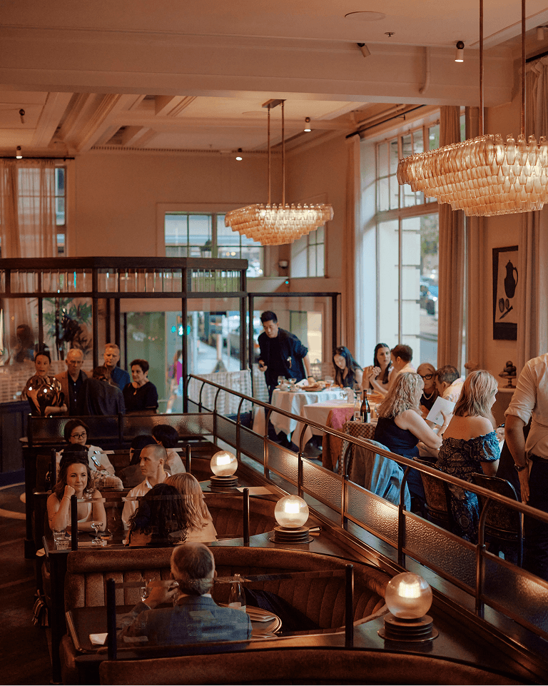 Elegant restaurant interior with chandeliers, patrons dining at booths and tables, warm lighting, and a server attending to a group in the background.
