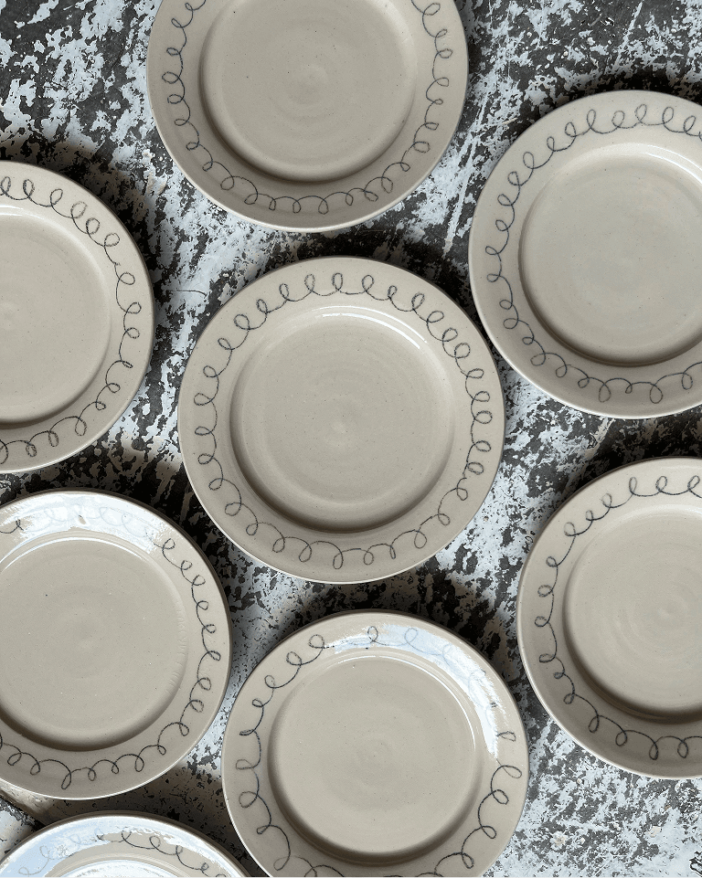 Beige ceramic plates with looped swirl patterns on the rims arranged on a textured, paint-splattered surface.