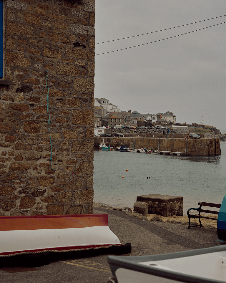 A stone wall with a blue rope, overturned boat, and a view of a calm harbor with moored boats, buildings, and overcast sky.