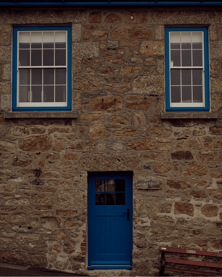 Stone building with two blue-framed windows, a blue door, and a sign reading "Wharf House." A wooden bench is in front of the wall.