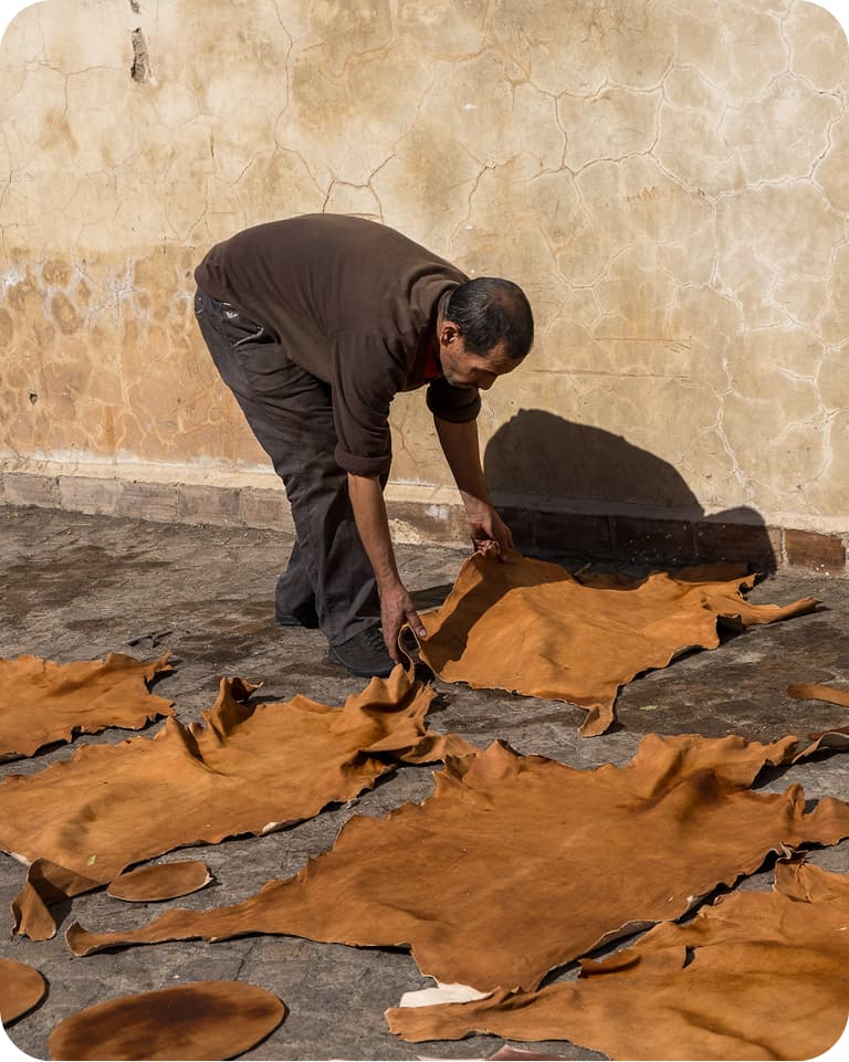 A person arranges brown leather hides on the ground against a textured wall under sunlight.