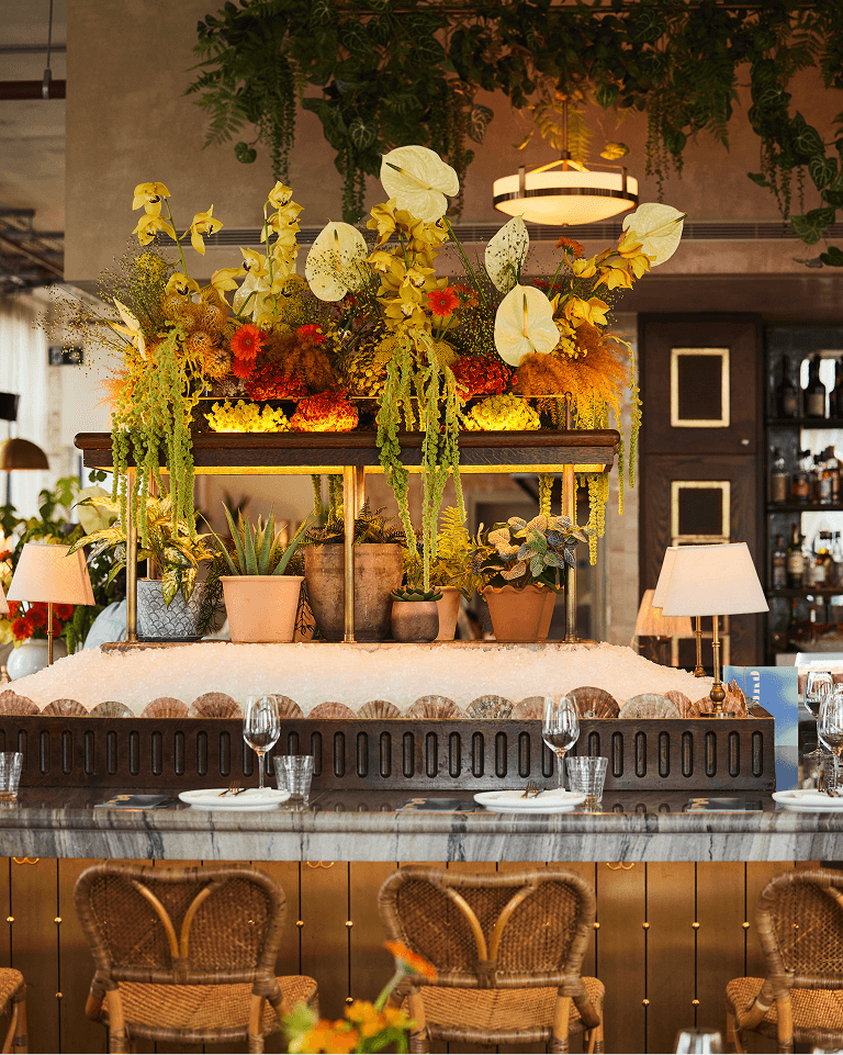 Elegant restaurant bar with lush floral arrangement, wicker chairs, and neatly set tables with wine glasses, creating a cozy and vibrant atmosphere.