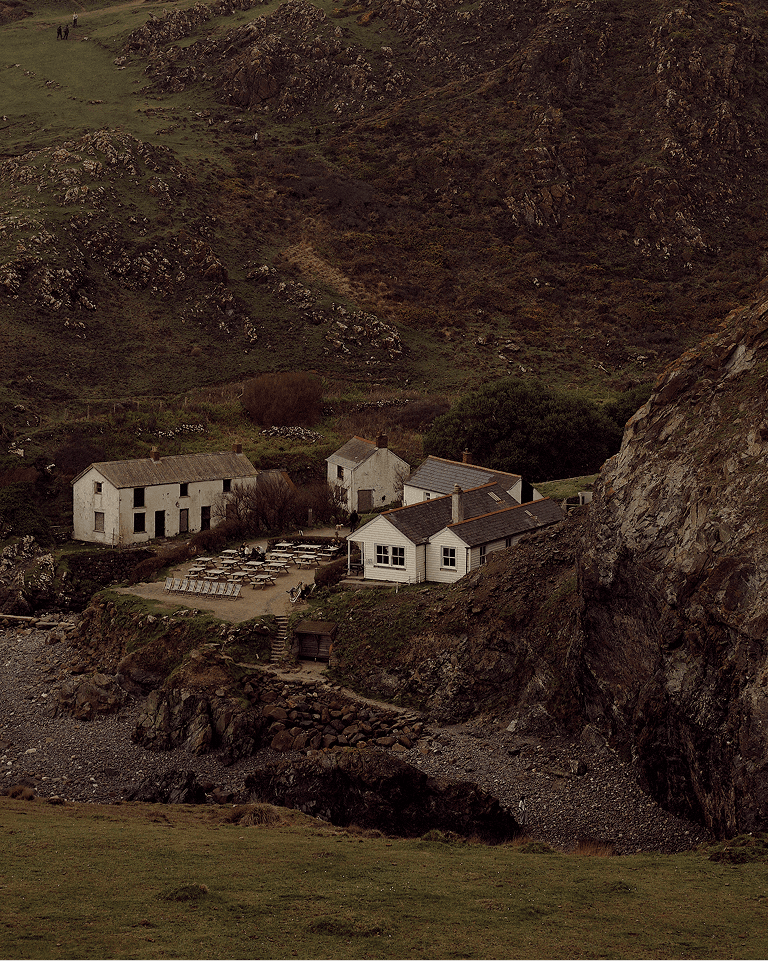 A cluster of small cottages nestled among rocky hills and a green landscape, with picnic tables and a path leading down to a rocky shoreline.