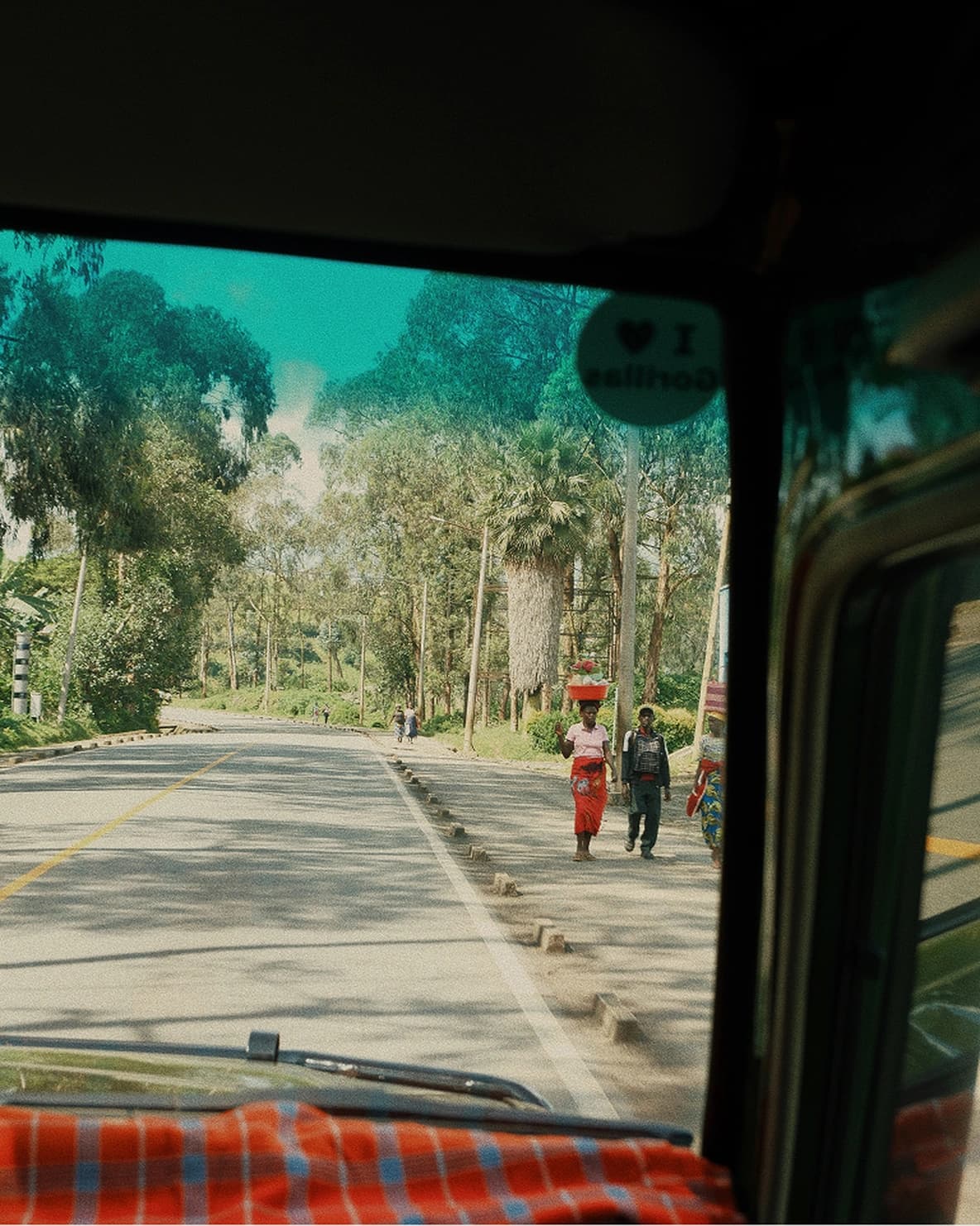 View through a vehicle windshield of a rural road, with people walking along the roadside. Trees and lush greenery line the road under a blue sky.