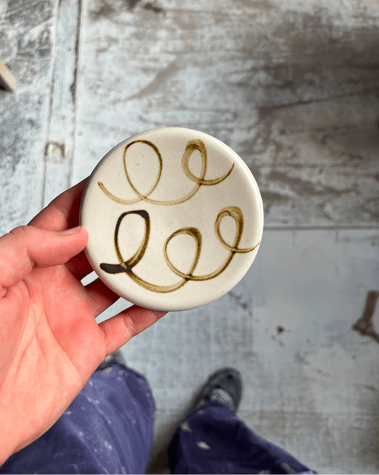A hand holds a round white ceramic tile with swirling brown line designs, set against a textured grey floor.