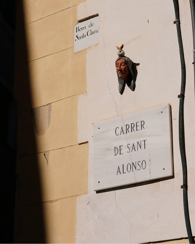 A street corner with signs for "Carrer de Sant Alonso" and "Born de Santa Clara," featuring a decorative head sculpture wearing a hat.
