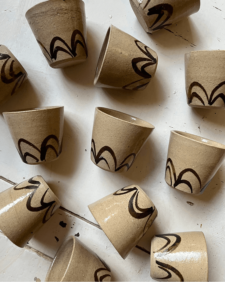 Twelve brown ceramic cups with dark abstract patterns are scattered on a white wooden surface.