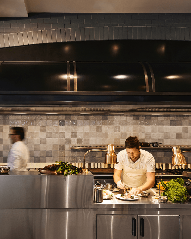 Chef plating dishes in a modern kitchen with a tiled backsplash, stainless steel surfaces, and various ingredients. Another person moves in the background.