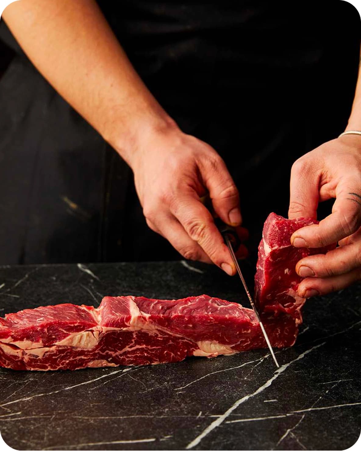 Hands slicing a raw strip of marbled beef with a knife on a dark marble countertop.