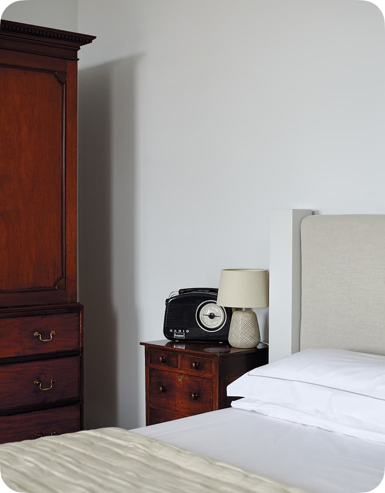 Bedroom corner with a wooden wardrobe, a bedside table with a vintage radio and lamp, and a neatly made bed with white linens.