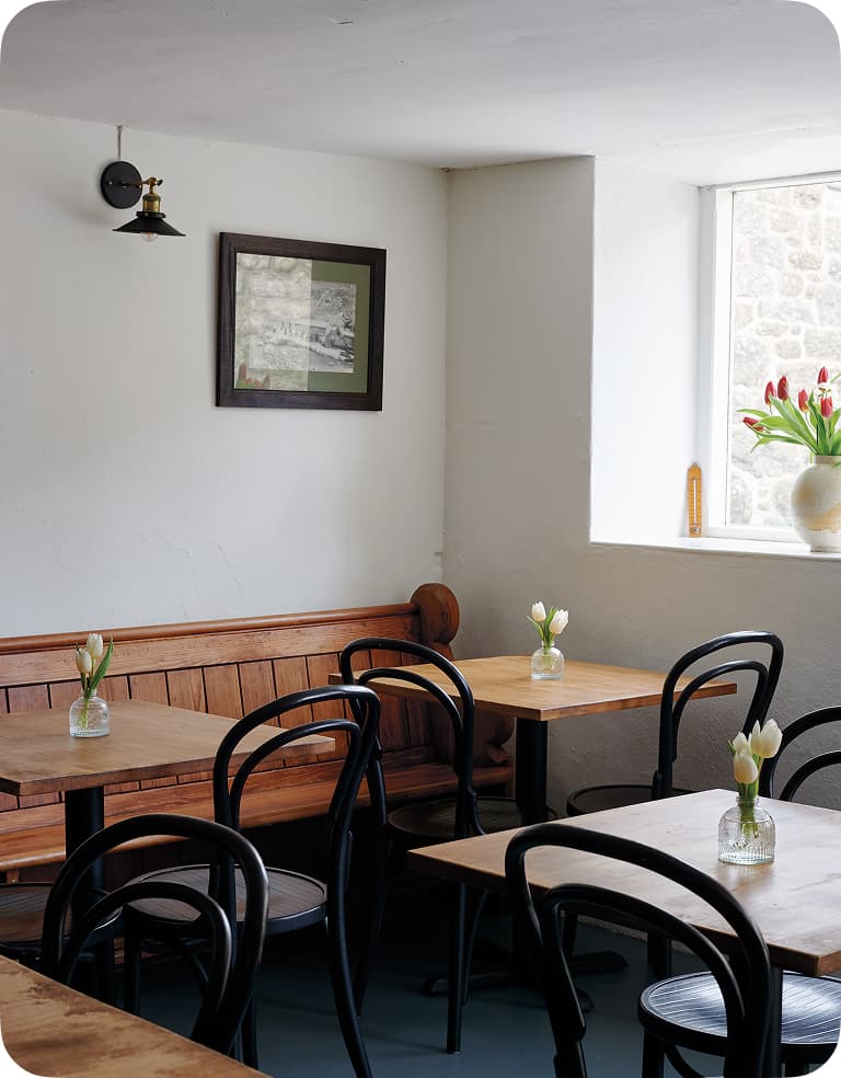 Cozy cafe interior with wooden tables and chairs, small vases with tulips, a wall picture, and a window letting in natural light.