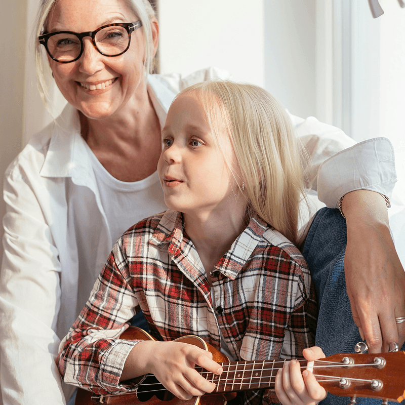 Elderly woman with glasses smiling, sitting beside a young child playing a ukulele, both in casual clothing near a bright window.