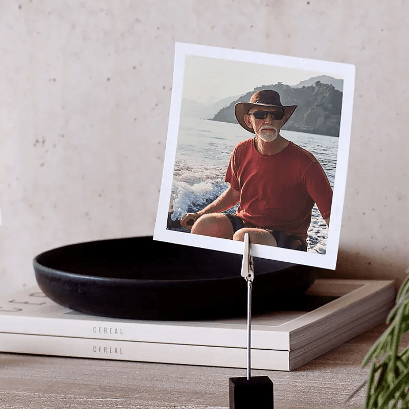 A photo of a man in a hat and sunglasses sitting by the sea, displayed on a clip stand.