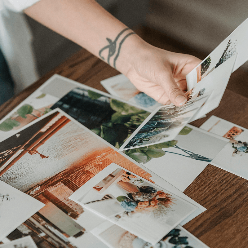 A person with a tattooed wrist sorts through a collection of colourful photographs spread out on a wooden table.