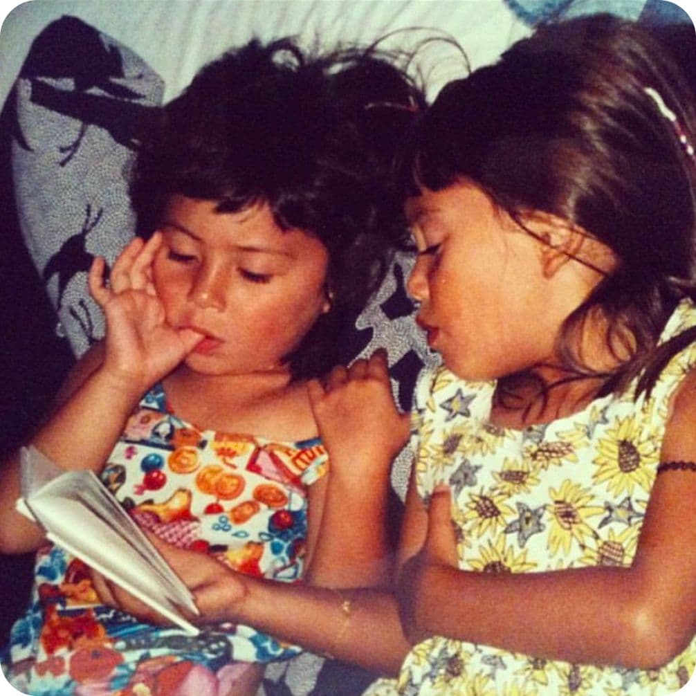 Two young girls lying on a bed, one reading a book while the other listens intently, wearing colorful dresses.