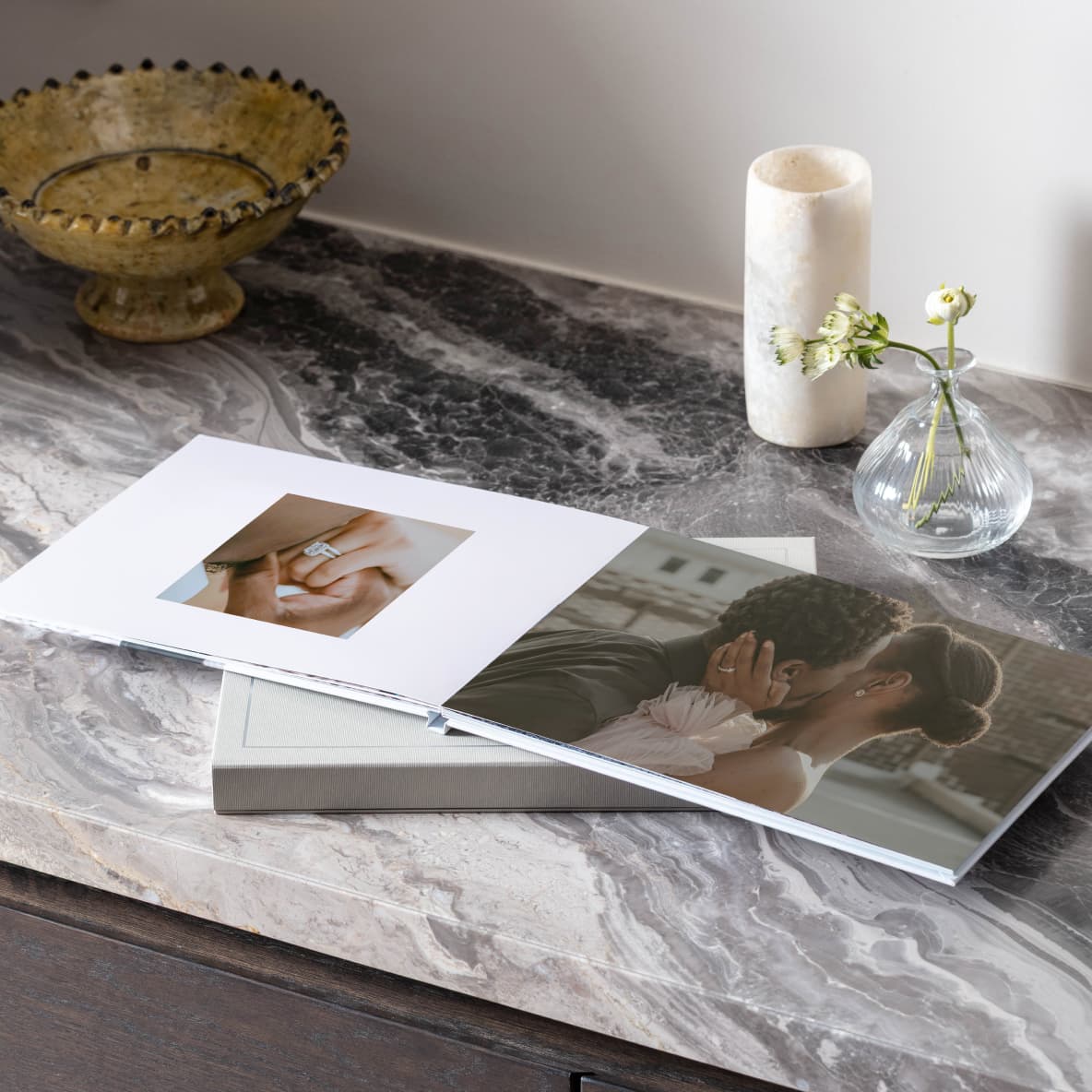An engagement photo book with layflat pages sits open on a marble counter, the photo book is open on a photo of a couple kissing and of the woman's engagement ring