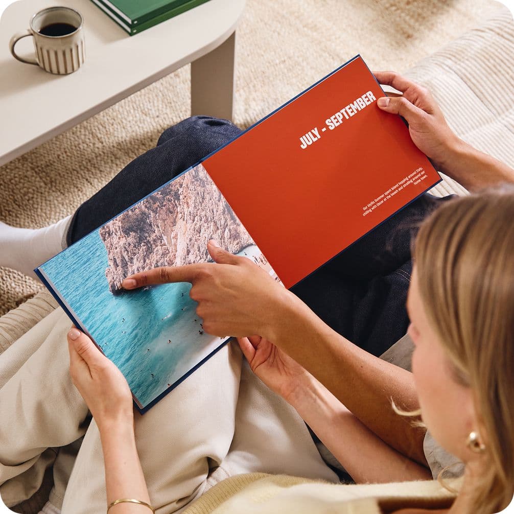 Two people sitting on a couch, looking at a photo book. One points to a page labeled "July - September" with a coastal image.