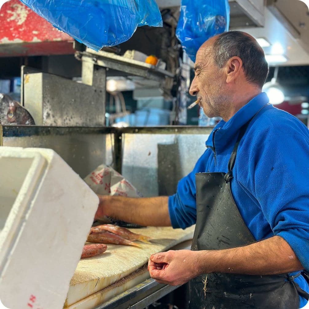 Man in a blue sweater and black apron cleaning fish at a market counter, with a cigarette in his mouth.