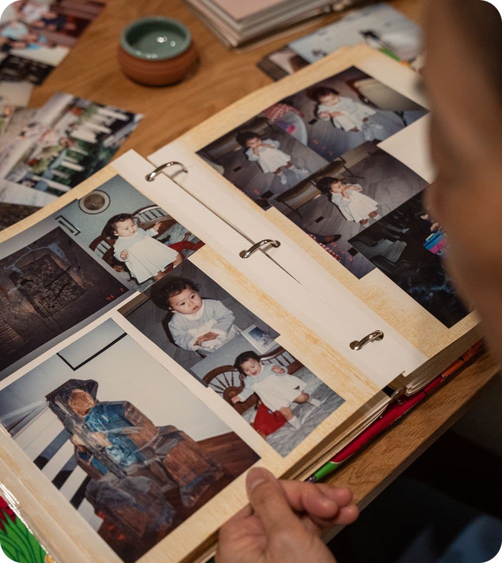 Person looking at a photo album with childhood pictures on a wooden table, surrounded by more photos and a small bowl.