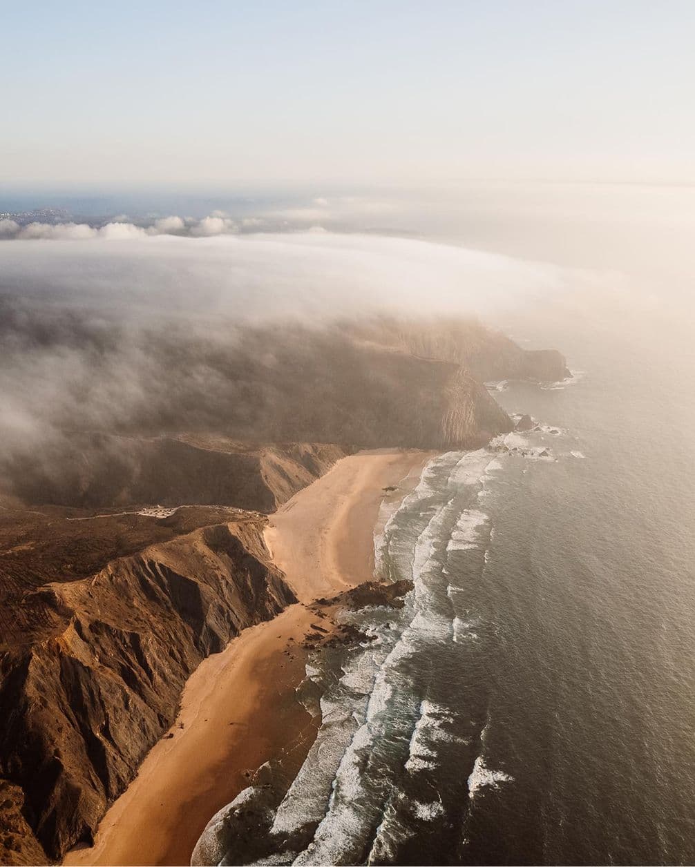 Aerial view of a rugged coastline with sandy beaches, cliffs, and waves under a cloudy sky, creating a serene and dramatic landscape.