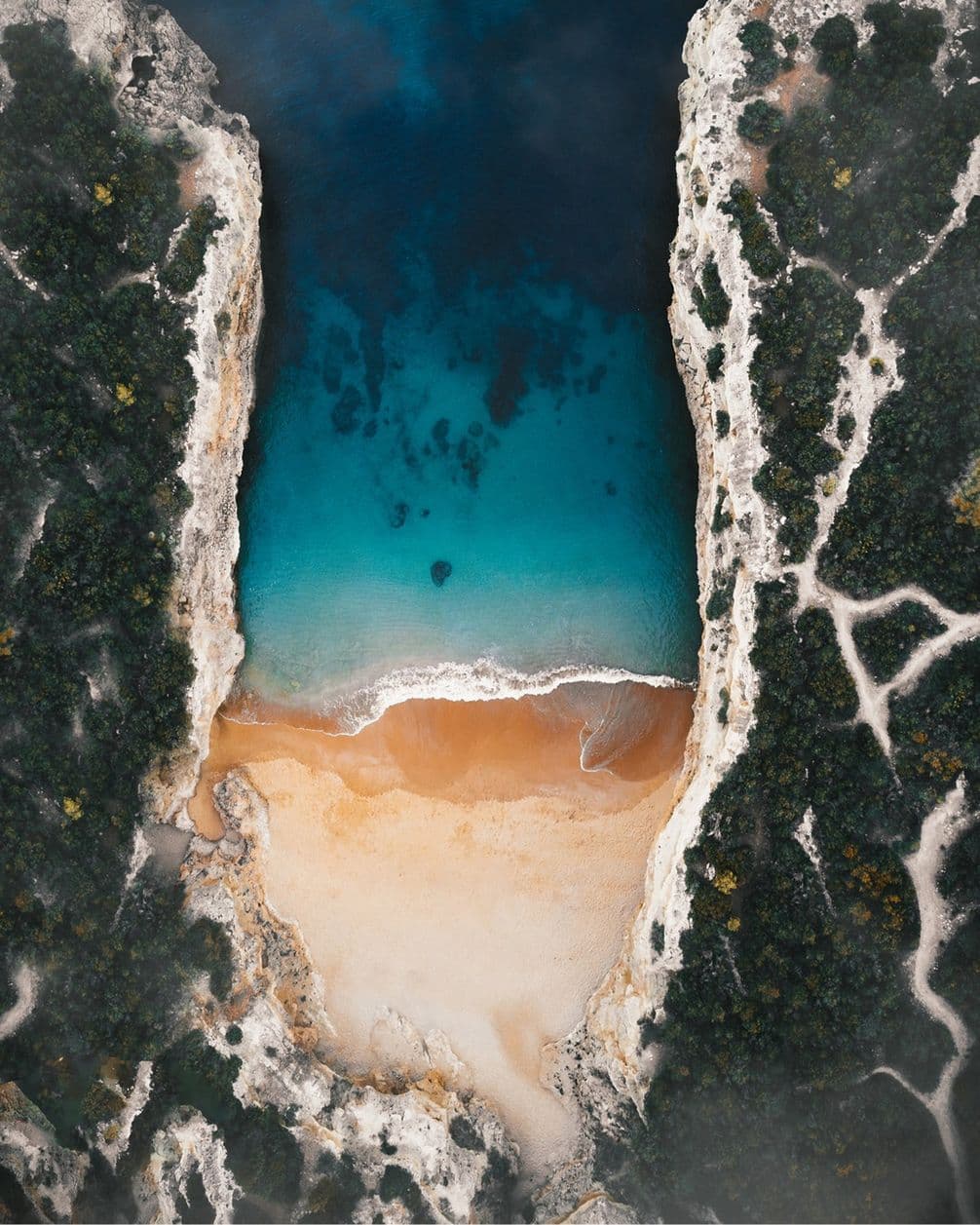 Aerial view of a secluded beach with turquoise water, surrounded by rocky cliffs and lush greenery, forming a narrow inlet.