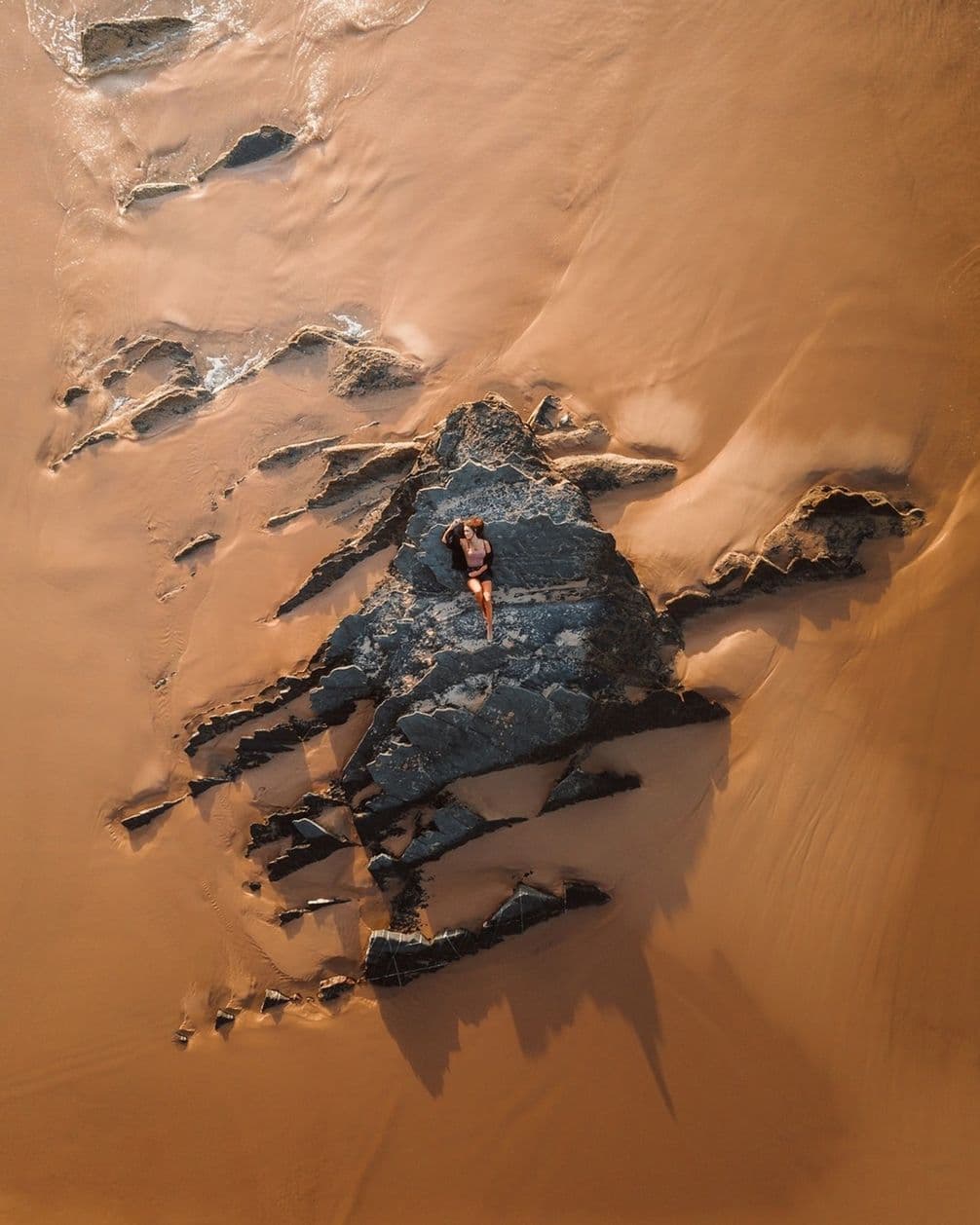 A person lies on dark rocks surrounded by smooth, wet sand on a beach, viewed from above. Shadows stretch across the sand.