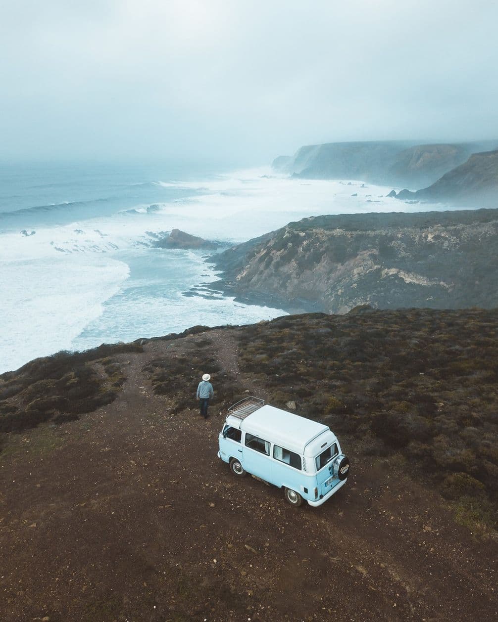 A person stands near a vintage van on a cliff overlooking a vast, cloudy seascape with rugged coastline and waves crashing below.