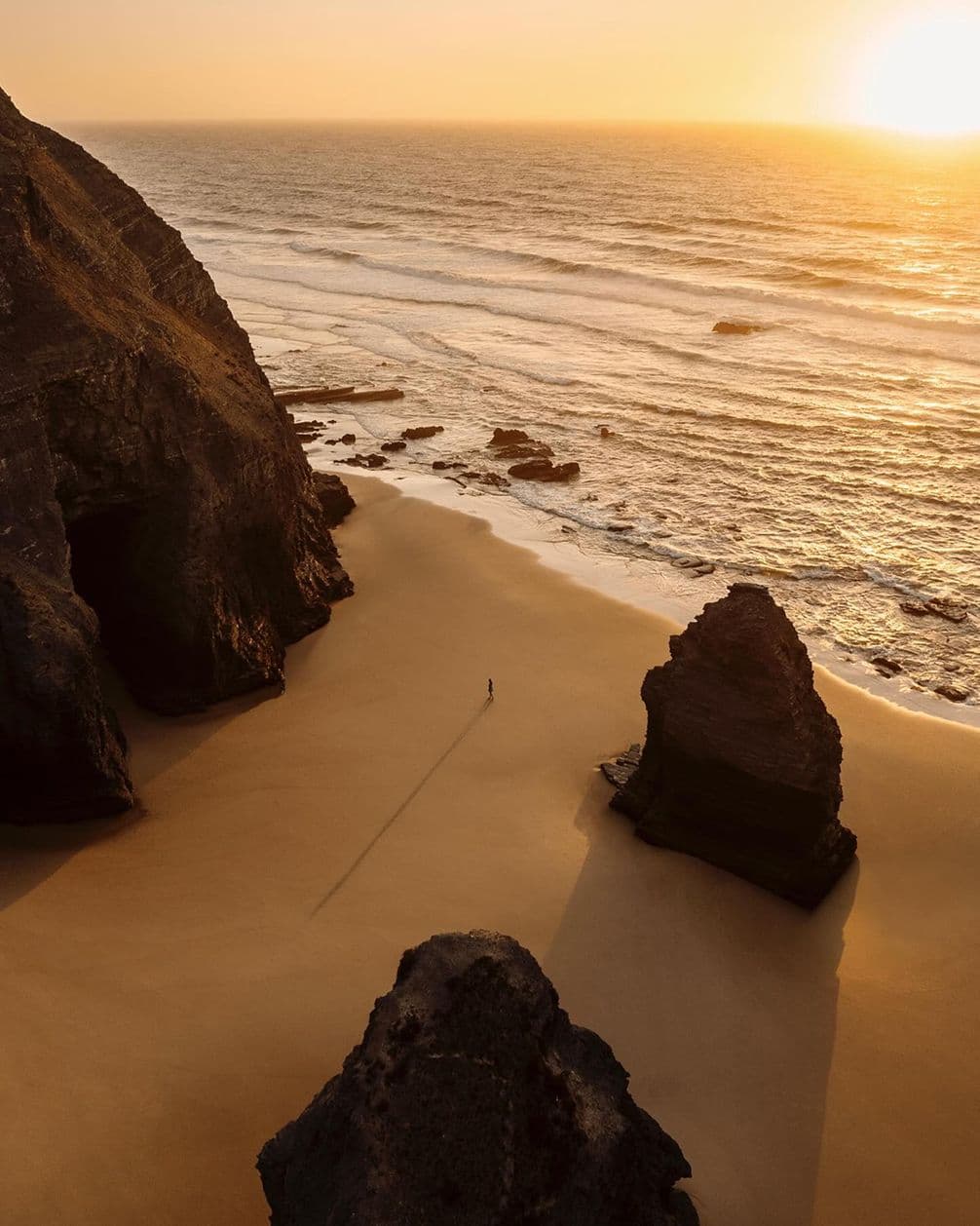 A person walks on a vast, sandy beach at sunset, surrounded by large rock formations and gentle ocean waves.