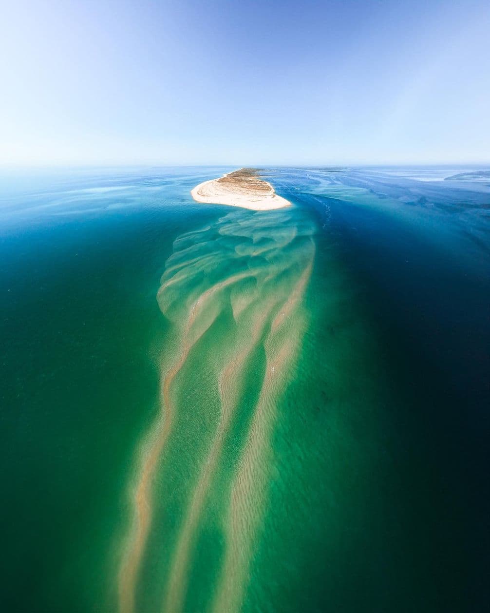 Aerial view of a sandy island surrounded by turquoise water, with sandbars creating wavy patterns beneath the surface.