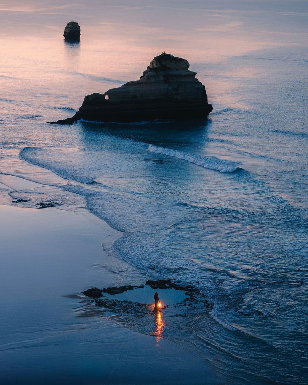 A person stands with a lit torch by the shoreline at dusk, with rock formations and gentle waves in the background.