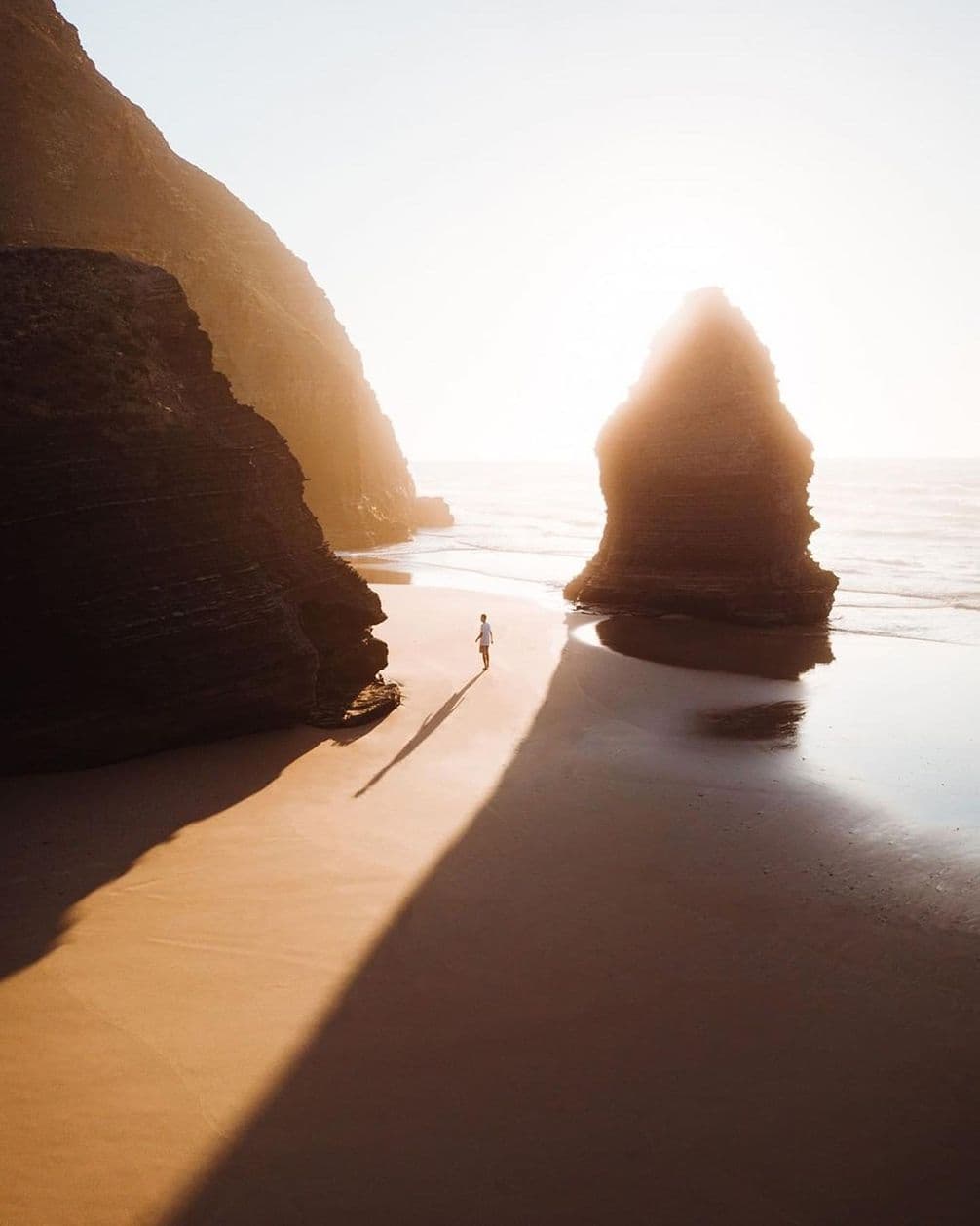 Person walking on a sunny beach between large rock formations, casting long shadows on the sand, with the ocean in the background.