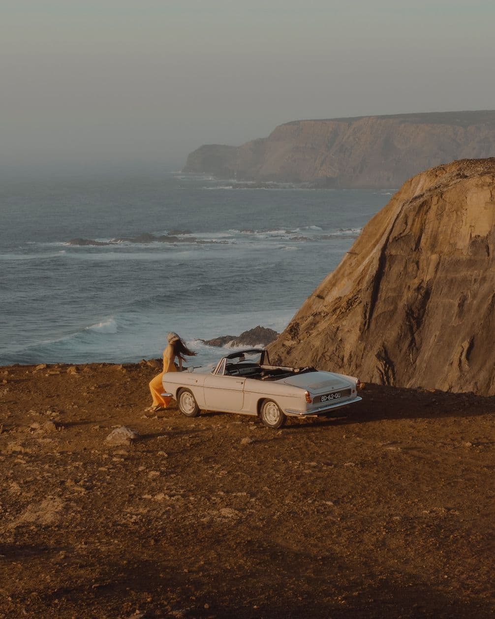 Woman in orange dress leans on a white convertible parked on a cliffside, overlooking the ocean and rocky coastline under a clear sky.