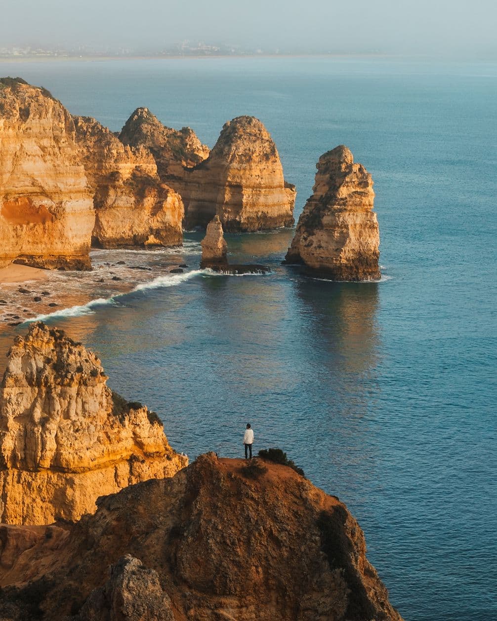 Person standing on a cliff overlooking the ocean with rugged rock formations and calm blue water under a clear sky.