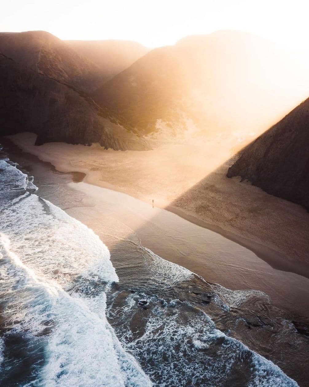 Aerial view of a sunlit beach with waves, surrounded by rocky cliffs. A person stands alone on the sand, bathed in golden light.