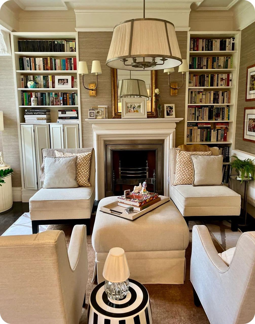 Cozy living room with armchairs, a fireplace, bookshelves, and a central ottoman. A large pendant light hangs overhead.