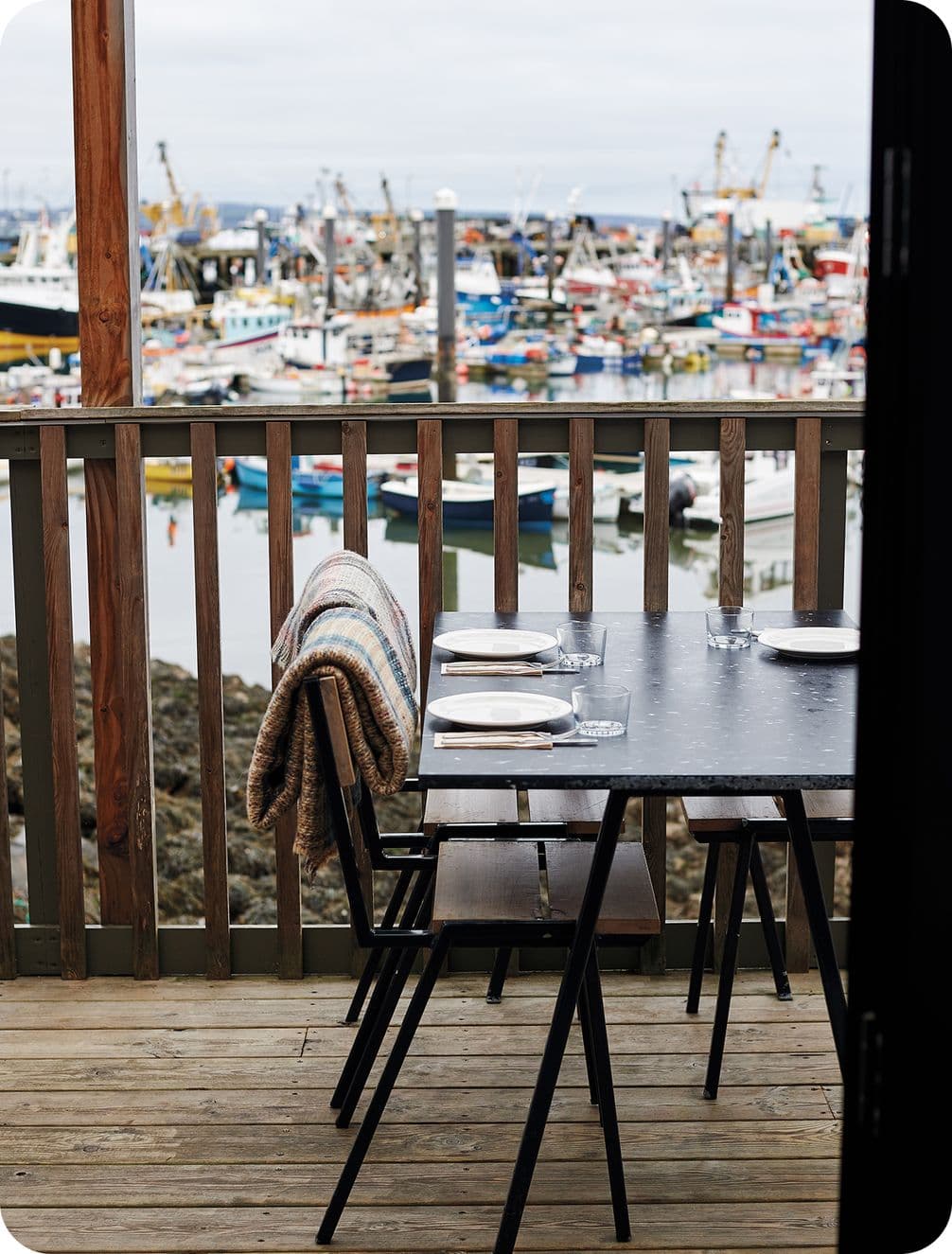 Outdoor dining table with plates and glasses on a wooden deck, overlooking a busy harbor filled with colorful boats.
