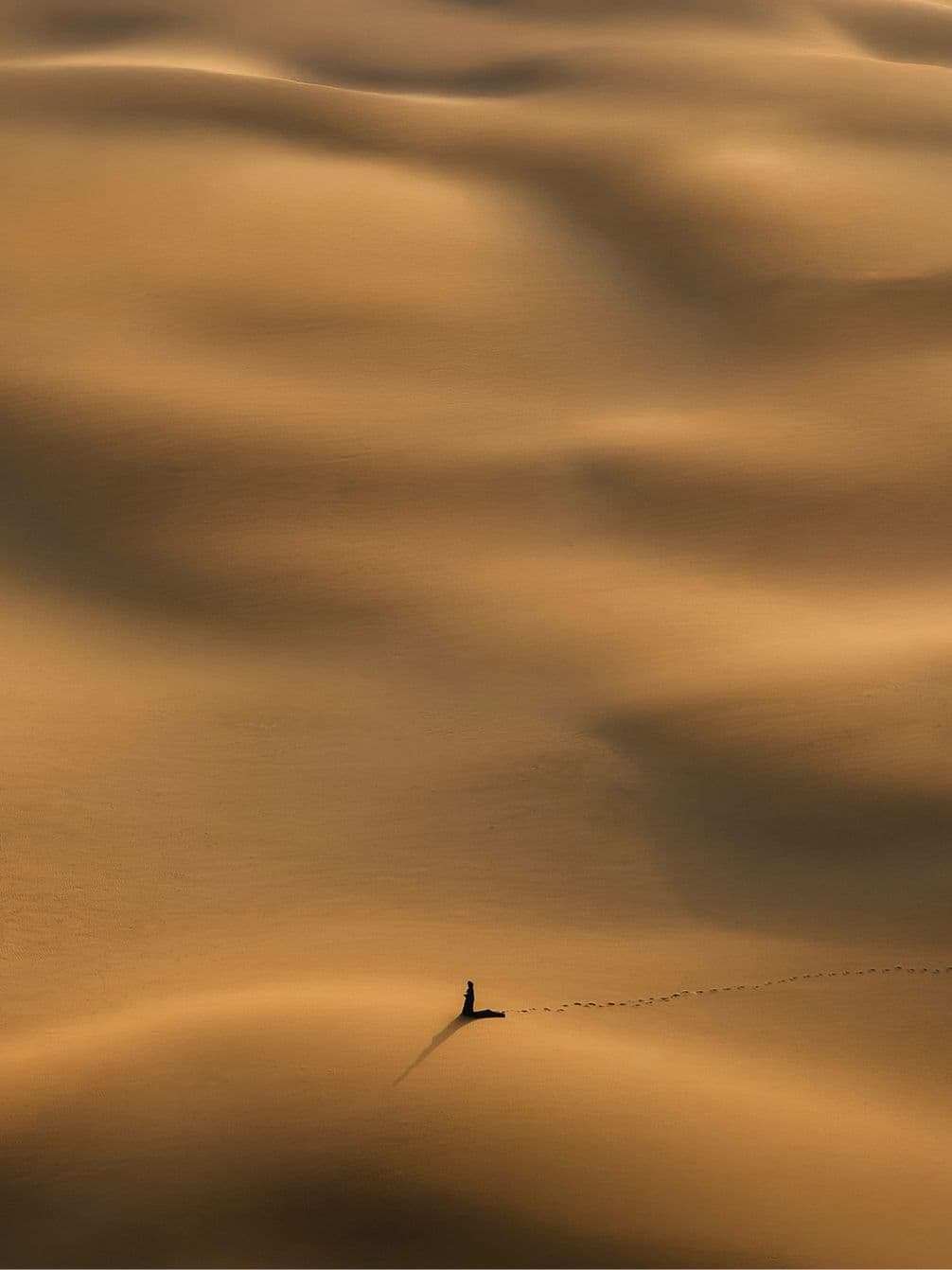 Drone photo of a lone person walking across vast, smooth sand dunes, leaving a trail of footprints under the warm, golden light.