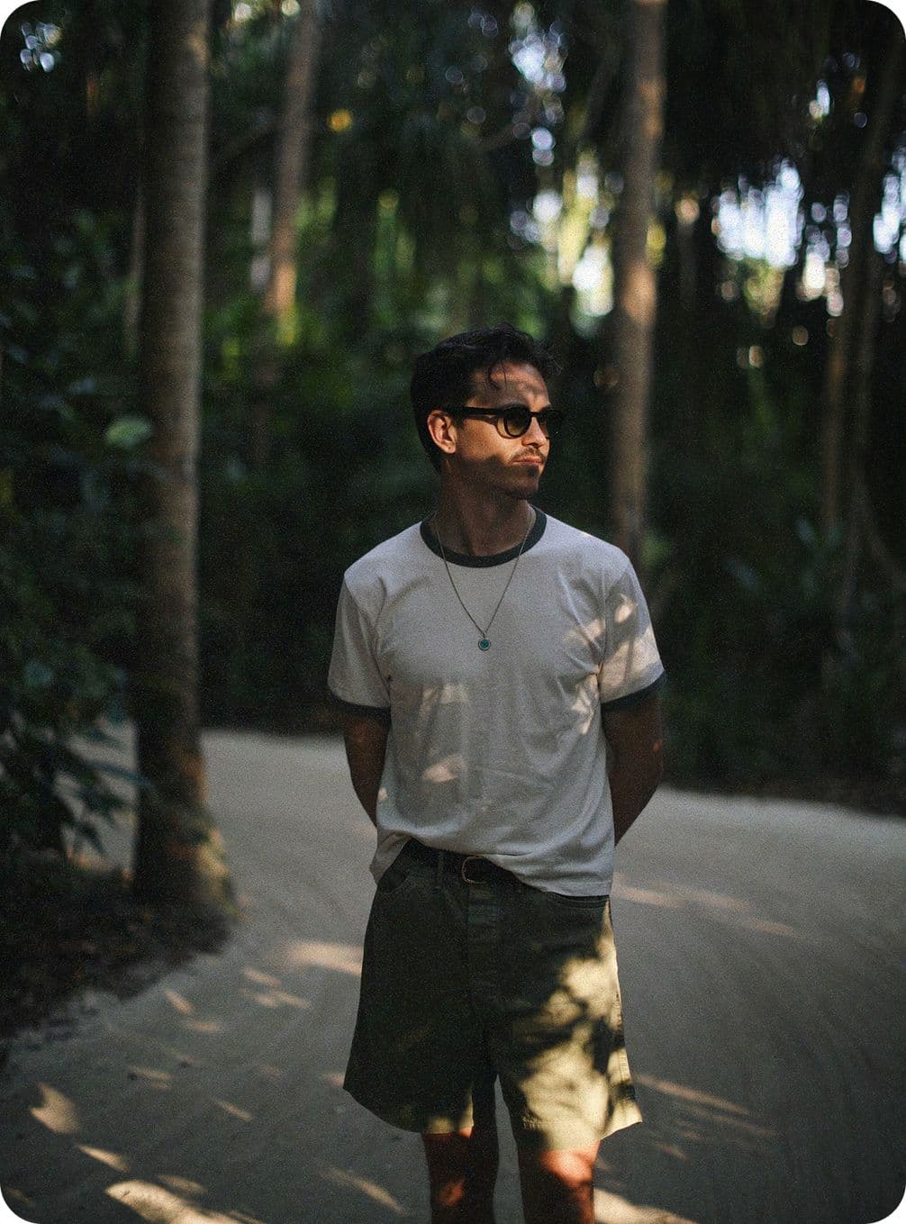 A person in a white shirt and shorts stands on a sunlit path in a forest, wearing sunglasses and a necklace.