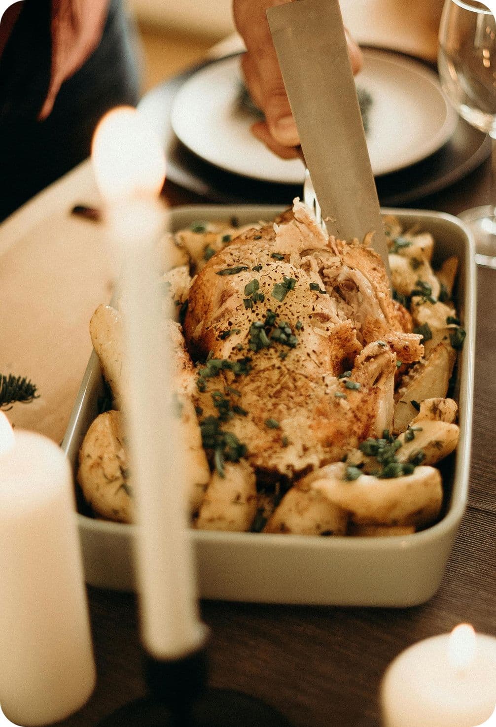 A person carving a roasted chicken garnished with herbs in a dish, surrounded by lit candles on a dining table.