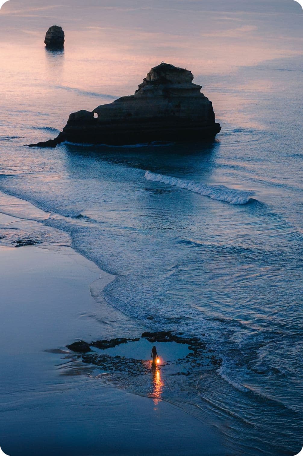 A person stands on a beach at sunset, with gentle waves and a large rock formation in the sea, reflecting warm light on the water.