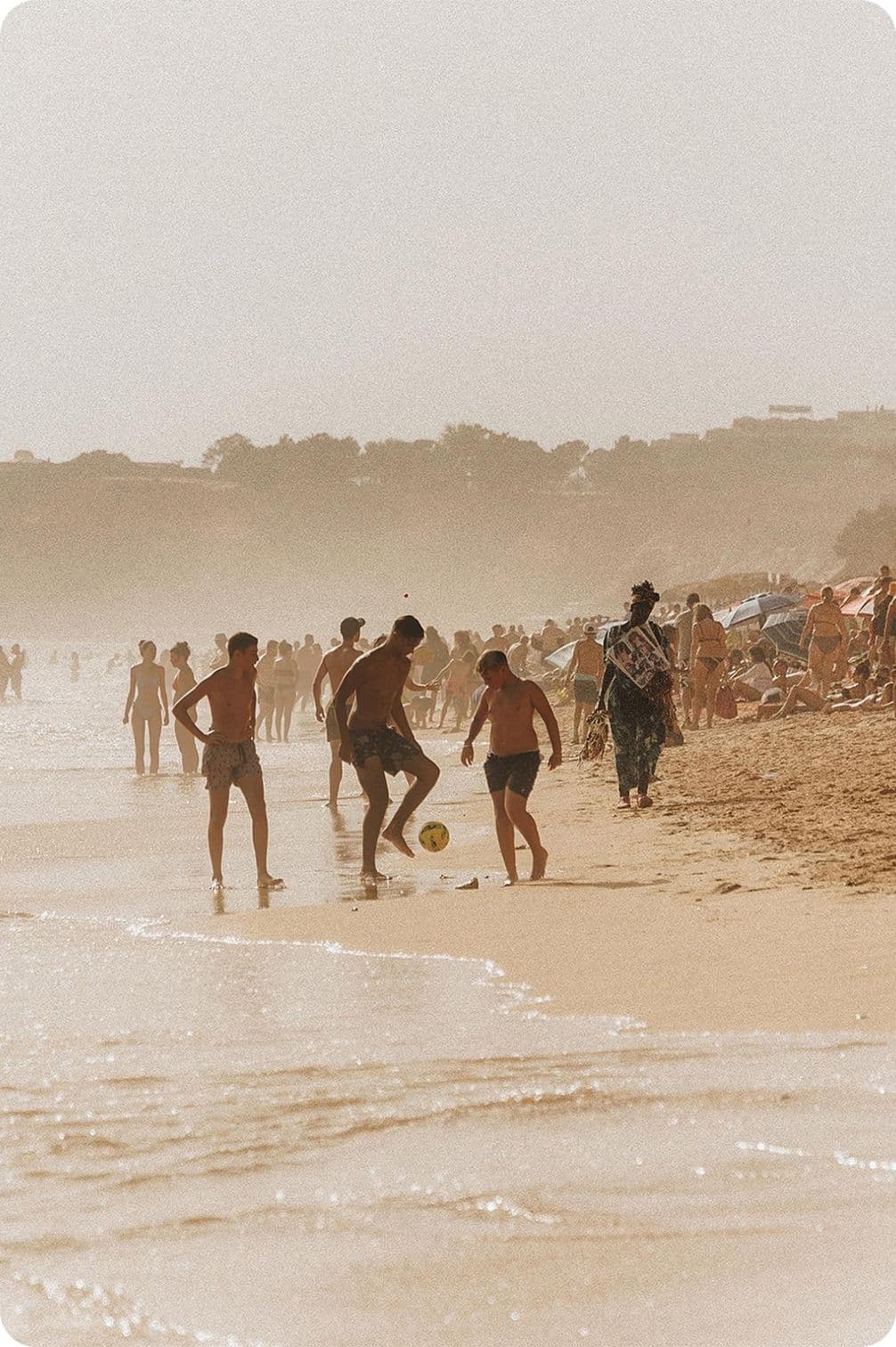 People playing soccer on a crowded sandy beach, with waves and umbrellas in the background.