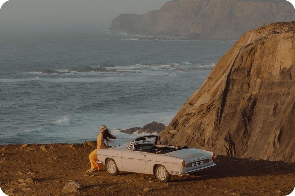 Person sitting on a white convertible by a cliff, overlooking the ocean with rocky hills in the background.