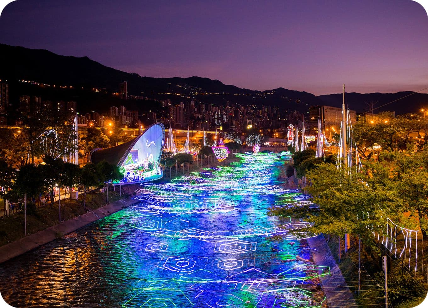A vibrant cityscape at dusk with colorful lights illuminating a river, surrounded by trees and buildings against a mountainous backdrop.