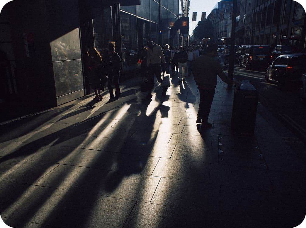 People walking on a city sidewalk at sunset, casting long shadows. The street is lined with buildings and cars.