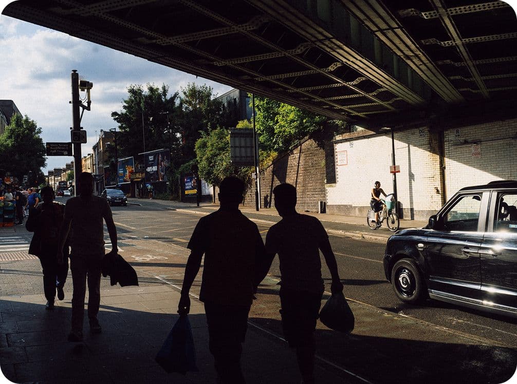 Silhouetted pedestrians walk under a bridge on a sunny street, with a cyclist and a black taxi visible in the background.